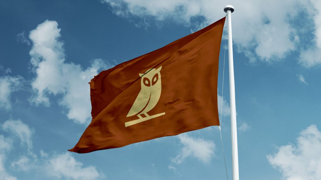 Rust-colored flag featuring the owl logo of Manifest Building, a Montecito-based landscape design firm, waving against a blue sky. The minimalist owl symbolizes clarity, vision, and refined craftsmanship in luxury outdoor design across Santa Barbara.
